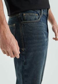 Ciemo niebieskie dżinsy z pomarańczowym przeszyciem, slim fit, widoczne kieszenie boczne i tylne, noszone z czarną koszulą.