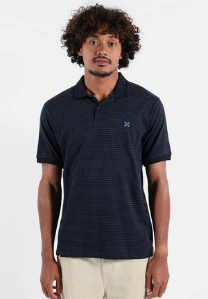 Homme aux cheveux bouclés portant un polo bleu marine à manches courtes et un pantalon beige, debout devant un fond blanc uni.