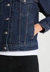 Persona che indossa una giacca in denim blu scuro con bottoni metallici, mano riposta nella tasca destra, visibile una camicia bianca e pantaloni neri.