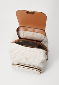 Sac à dos avec un rabat supérieur en cuir tan et des accessoires en or. Le corps en tissu crème présente un motif de logo répétitif. Comprend des doubles fermetures éclair et des poches intérieures.