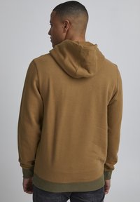 Felpa marrone con cappuccio, texture lavorata a maglia e polsini e orlo a coste. Presenta un accento verde stratificato lungo l'orlo inferiore.
