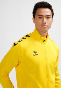 Gul zip-up jacka i slät tyg med hög krage, svarta accenter på axlarna och en liten logotyp på bröstet.