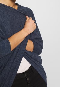 Person in einem dunkelblauen Strick-Wickel mit einem Ellbogen-Ausschnitt, über einem weißen Oberteil und schwarzen Hosen mit mehreren Knöpfen.