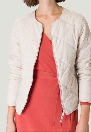 Vrouw draagt een licht beige doorgestikte jas over een koraalroze jurk met V-hals, waarbij de jas gedeeltelijk is opengerit zodat details van de jurk zichtbaar zijn.