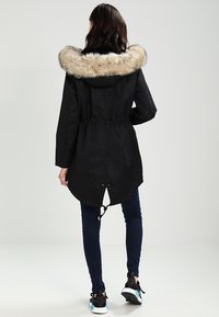 Svart parkas med en huva fodrad med fuskpäls, dragsko vid midjan och rundad nederkant. Bärs över mörka jeans och svarta sneakers med blå detaljer.