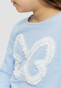 Lichtblauwe sweatshirt met een wit gerimpeld vlinderontwerp. Zachte stof met een ronde halslijn en lange mouwen.