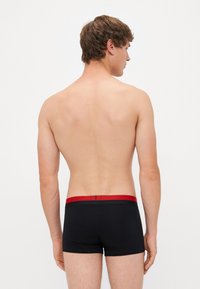 Schwarze Boxershorts mit rotem elastischem Bund; glatte Textur, enganliegendes Design und Oberschenkel-lange Länge. Von hinten betrachtet.