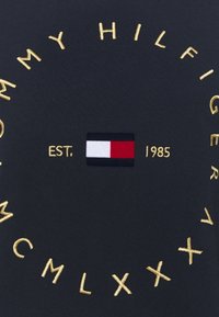 Tela negra con texto bordado en oro: "TOMMY HILFIGER," "EST. 1985," y números romanos, además de un pequeño logotipo rectangular en rojo, blanco y azul.