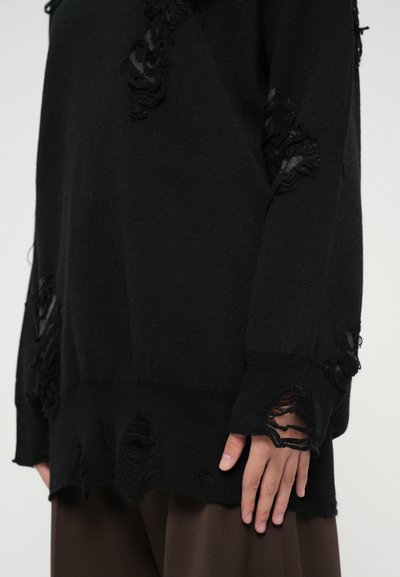Black knit sweater featuring distressed detailing with frayed edges. Long sleeves and an oversized fit, paired with brown trousers.