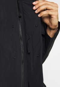 Veste noire avec une fermeture éclair intégrale, des boutons-pression et une poche intérieure visible avec un onglet de tirage. Le matériau semble léger et lisse.