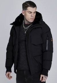 Black puffer jacket with a faux fur-lined hood, multiple front pockets, and a logo patch on the sleeve. Worn over a dark shirt.