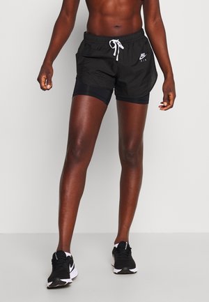 Sportliche Person trägt schwarze Nike-Shorts mit weißem Kordelzug und schwarze Laufschuhe, steht vor schlichtem Hintergrund.