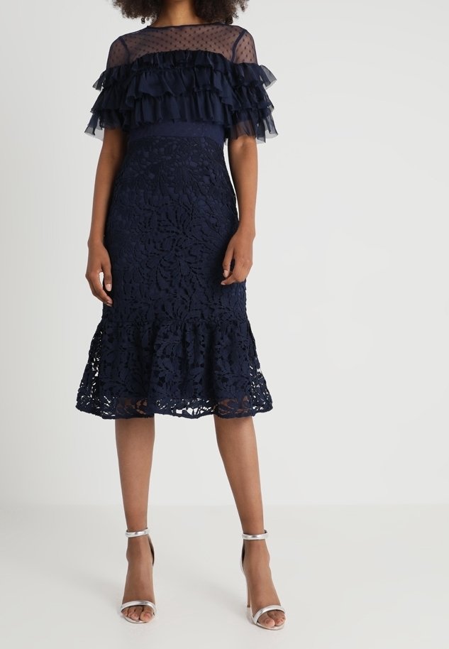 Robe en dentelle bleu marine avec manches transparentes à volants superposés et ourlet évasé. Talons argentés à brides complètent le look. Motif floral texturé sur toute la robe.