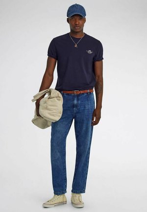 Uomo con t-shirt blu navy, jeans blu, scarpe da ginnastica beige, cappellino blu, che tiene una giacca beige nella mano destra su sfondo uniforme.