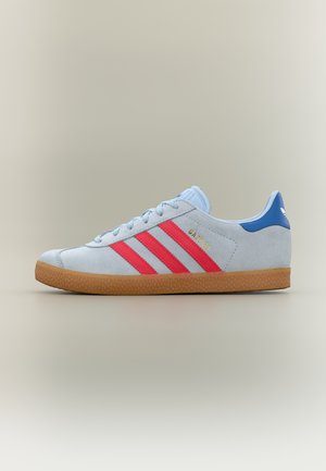 Hellblauer Adidas Gazelle Sneaker mit roten Streifen, Kaugummisohle und blauem Fersenlaschen vor neutralem Hintergrund.