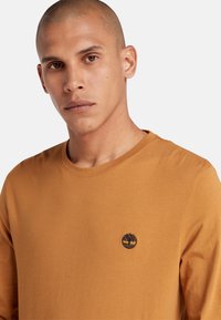 Langarmshirt in amberkleur, gemaakt van zachte stof, met een ronde hals en een klein donker logo op de borst.