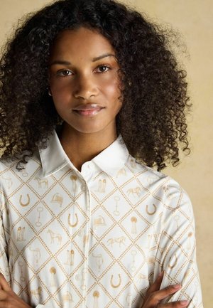 Junge Frau mit lockigem Haar trägt ein weißes Hemd mit Knöpfen, das mit hellbraunen Reitsport-Symbolen auf beigem Hintergrund gemustert ist.