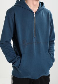 Niebieska bluza z kapturem na zamek błyskawiczny, z przednią kieszenią i czarną grafiką z logo na piersi, wykonana z miękkiego materiału.