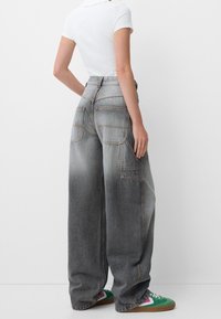 Szerokie szare dżinsy z jaśniejszymi wstawkami, wyraźnym przeszyciem i dwiema tylnymi kieszeniami, noszone z zielonymi trampkami.