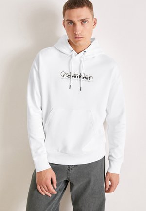 Sudadera de algodón blanca con un bolsillo canguro en la parte frontal, cordones y un logo en negro y marrón en el pecho. Textura suave, corte relajado.