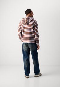 Mann som står med ryggen til, iført en brun og hvit stripete hettegenser, falmede blå jeans og hvite joggesko med brune striper, mot en hvit bakgrunn.