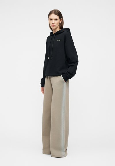 Γυναίκα που στέκεται φορώντας μαύρο φούτερ με κουκούλα και λογότυπο στο στήθος και μπεζ παντελόνι φαρδιάς γραμμής με ανοιχτές ρίγες στο πλάι, με τα χέρια στις τσέπες.