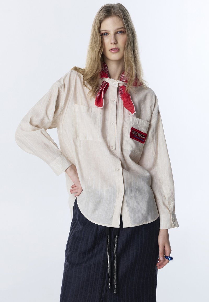 Junge Frau trägt ein beiges Hemd mit roten Aufnähern und ein rotes Halstuch, posiert mit einer Hand in der Hüfte vor weißem Hintergrund.