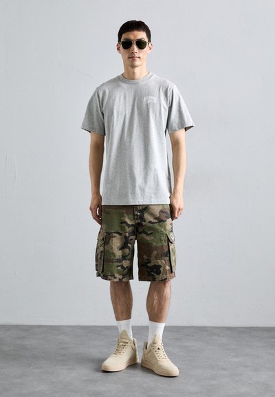 Tânăr stând, purtând ochelari de soare negri, un tricou gri, pantaloni scurți cargo cu camuflaj, șosete albe și adidași bej, pe un podea gri.