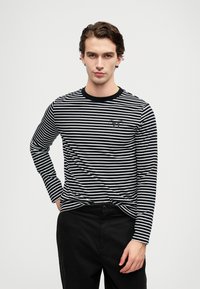 Fekete-fehér csíkos, hosszú ujjú póló fekete kereknyakkal és kis logóval a mellkason, fekete nadrággal párosítva.