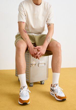 Person med tatoveringer iført beige Nike T-shirt, olivengrønne shorts, hvide Nike Air Max sneakers og hvide sokker, siddende på en metalopbevaringskasse på en gul gulv.