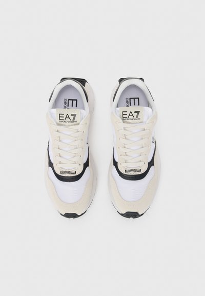 Des chaussures de sport beiges et blanches présentant des matériaux en mesh et en suède, des accents noirs, une languette rembourrée, et un logo sur la languette et le côté.