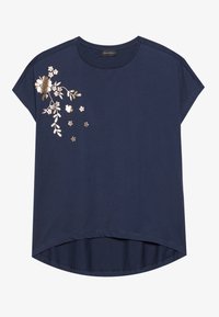 Blouse bleu marine à manches courtes avec un col rond et une broderie florale dorée sur l'épaule gauche et la zone de la poitrine.