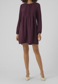 Langärmliges, dunkelviolettes Kleid mit gefaltetem Vorderteil, lockerer Passform, rundem Ausschnitt und Knopfinfos an der Oberseite; glatte Stofftextur.