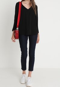 Černé dlouhé triko s výstřihem do V, spárované s tmavými džíny a bílými teniskami. Součástí je malá červená kulatá kabelka přes rameno s červeným popruhem.