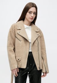 Jonge vrouw met lang bruin haar, gekleed in een beige shearling jas over een wit shirt en zwarte leren broek, staand tegen een effen achtergrond.