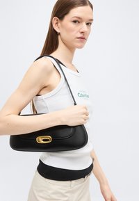 Γυναίκα με λευκό αμάνικο τοπ κρατάει μαύρη δερμάτινη τσάντα ώμου με χρυσό κούμπωμα, μπροστά σε απλό φωτεινό φόντο.