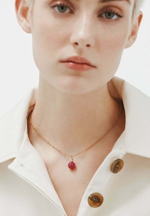 Collar de oro con un pequeño colgante redondo rosa, llevado sobre una camisa blanca con un cuello estructurado y acentos de grandes botones marrones.