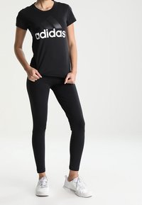 Černé tričko Adidas s velkým bílým logem, v kombinaci s černými legínami. Model má na sobě bílé sportovní boty. Hladká tkanina a přiléhavý design.