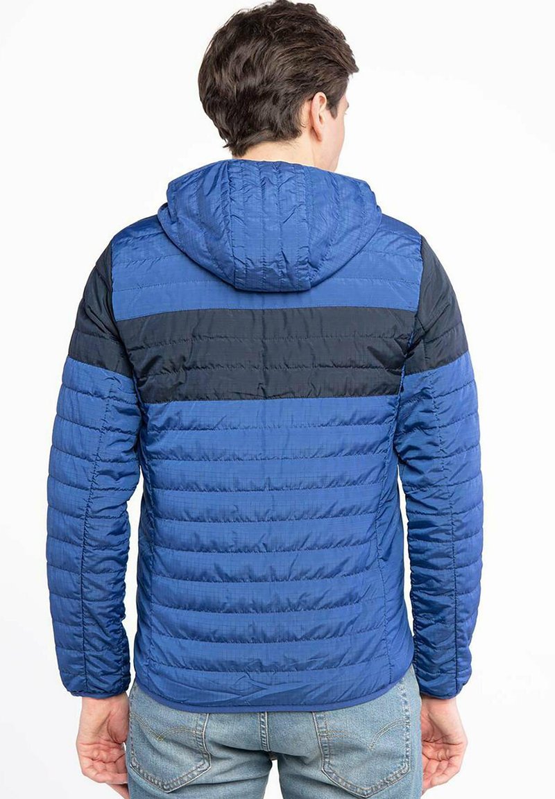 Mann trägt eine blaue Steppjacke mit schwarzem Horizontalstreifen und Kapuze, steht mit dem Rücken zur Kamera, und hat helle blaue Jeans an.