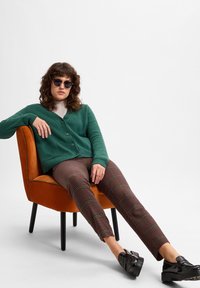Groene gebreide cardigan met een V-hals en knopen, gecombineerd met nauwsluitende pantalon met een multikleurig patroon. Zwarte plateau schoenen en ronde zonnebril.
