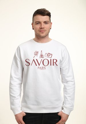 Mężczyzna w białej bluzie z ikonami róży, Wieży Eiffla i aparatu nad napisem „SAVOIR PARIS”, stojący na jednolitym tle.