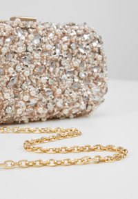 Collier chaîne en or attaché à une petite pochette ornée de cristaux, de perles et de perles sur une surface claire.