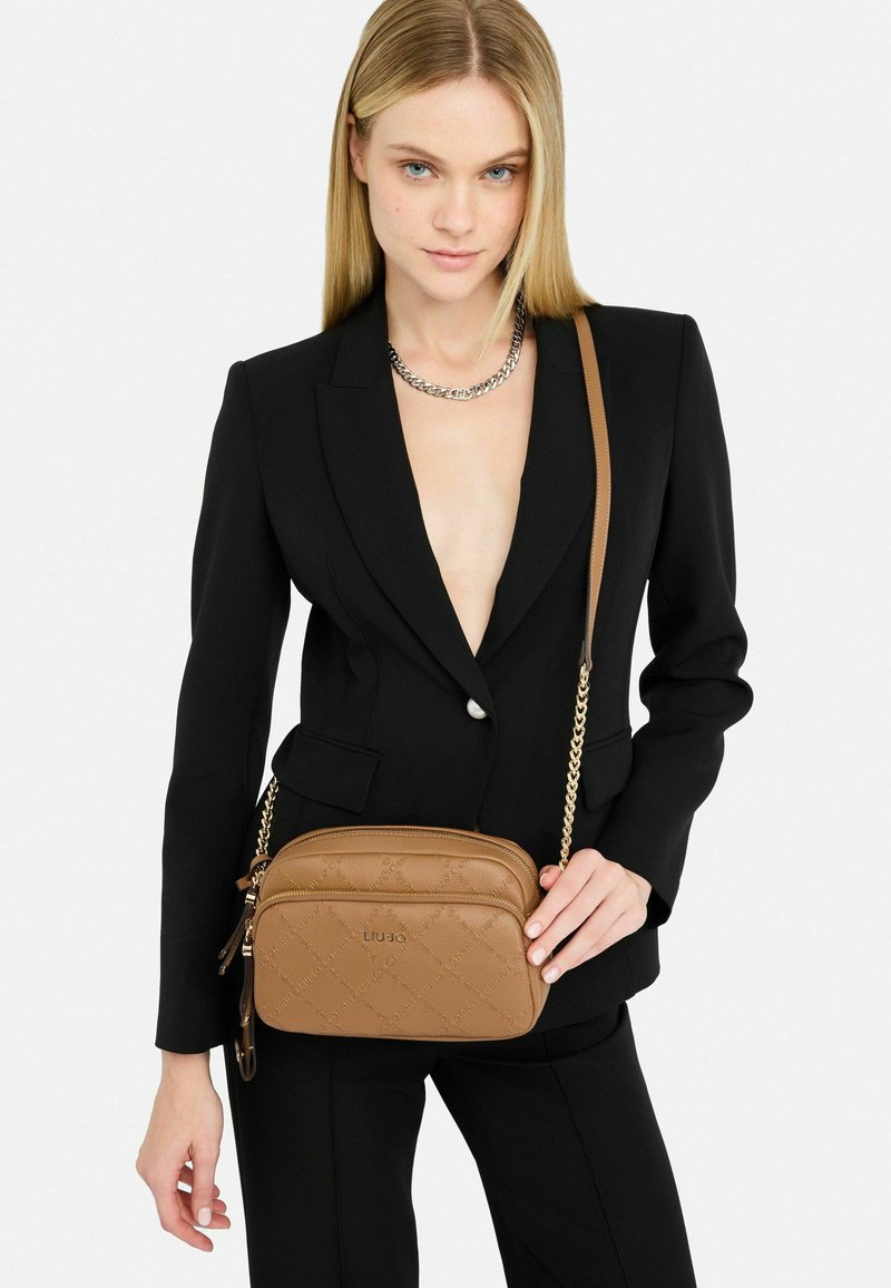 Hnědá kožená crossbody taška s embosovaným vzorem, dvojitými přihrádkami a zlatým řetězovým popruhem. Nositá s černým saka.