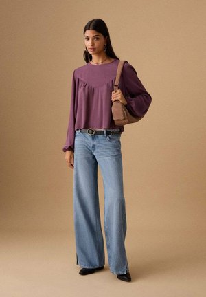 Long-sleeve purple top with ruffled detail, light blue wide-leg jeans, black shoes, and a small brown shoulder bag against a neutral background.