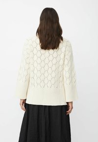 Gebreide crèmekleurige cardigan met een getextureerd opengewerkte patroon, lange mouwen en een ribbel boord. Draagt over een zwarte rok.