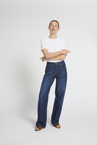 Žena stoji sa prekriženim rukama, nosi bijelu majicu kratkih rukava, široke plave traperice i smeđe sandale s otvorenim prstima, na jednostavnoj bijeloj pozadini.