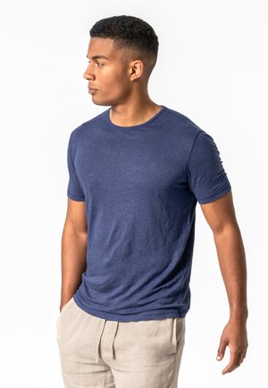 Junger Mann mit kurzen lockigen Haaren, der ein eng anliegendes marineblaues T-Shirt und beige Hosen trägt, schaut nach links, die Hände entspannt.