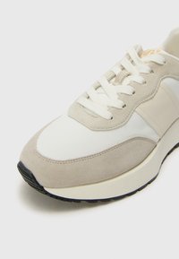 Sneaker grigio chiaro con una combinazione di tessuti morbidi e scamosciati, dotata di lacci bianchi, una suola imbottita e una suola esterna in gomma scura.