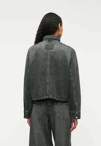 Szara dżinsowa kurtka o lekko krótkim kroju, z kołnierzem, zapięciem na guziki na mankietach oraz prostokątną czarną łatą na plecach.