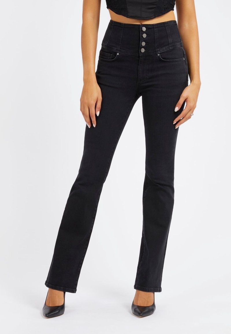Femme portant un jean noir taille haute avec quatre boutons, associé à des talons noirs pointus, debout devant un fond uni.